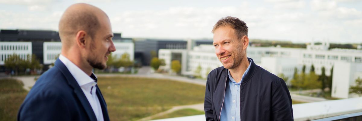 Zwei Mitarbeiter führen auf der Dachterrasse ein Gespräch und im Hintergrund ist der Innenbereich des Business Campus Unterschleißheim zu sehen