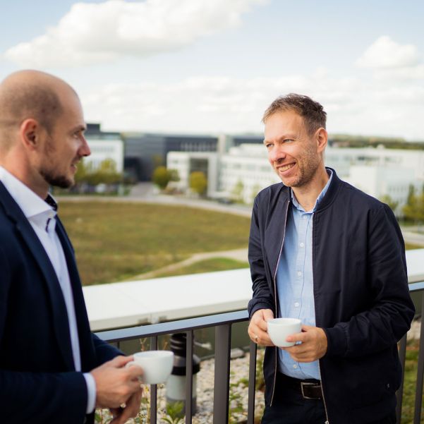 Zwei Mitarbeiter führen auf der Dachterrasse ein Gespräch und im Hintergrund ist der Innenbereich des Business Campus Unterschleißheim zu sehen